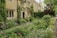 A charming stone house surrounded by a lush, well-maintained garden. The exterior features large windows, and the garden is filled with a variety of flowering plants and bushes. Ivy climbs up the house walls, adding to the cottage-like aesthetic. A pathway runs alongside the garden, lined with dense greenery.
