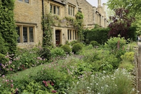 A charming stone house surrounded by a lush, well-maintained garden. The exterior features large windows, and the garden is filled with a variety of flowering plants and bushes. Ivy climbs up the house walls, adding to the cottage-like aesthetic. A pathway runs alongside the garden, lined with dense greenery.