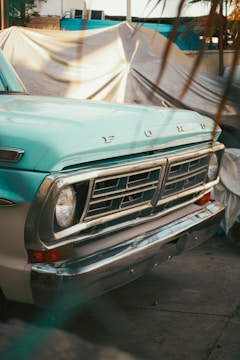 A vintage pick-up truck with a classic front grille and headlight design is parked in an outdoor setting. The truck has a pale turquoise color with chrome details. In the background, there is a tarp or cover draped over an object, suggesting a rustic or storage environment.