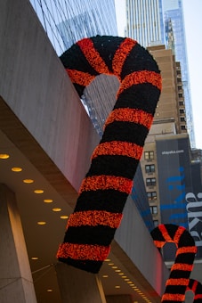 Large candy cane decorations with red and black stripes hang on the side of a building. They are part of festive decorations in an urban setting, surrounded by skyscrapers with glass and concrete facades. Several windows and some modern architectural details are visible, with the candy canes contrasting against the urban backdrop.