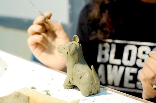 A person is sculpting a small clay figure resembling a dog. The hands are focused on shaping the clay with a tool. The person is wearing a black shirt with white lettering partially visible in the background.