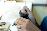 Hands are sculpting a small figure out of clay, using a fine tool to shape the details. The setting appears to be a well-lit workspace with a focus on craftsmanship.