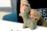 A person is sculpting a small clay figure of a dog with basic features and smooth texture. The focus is on the hands and the clay model, with tools used in sculpting visible in the background.