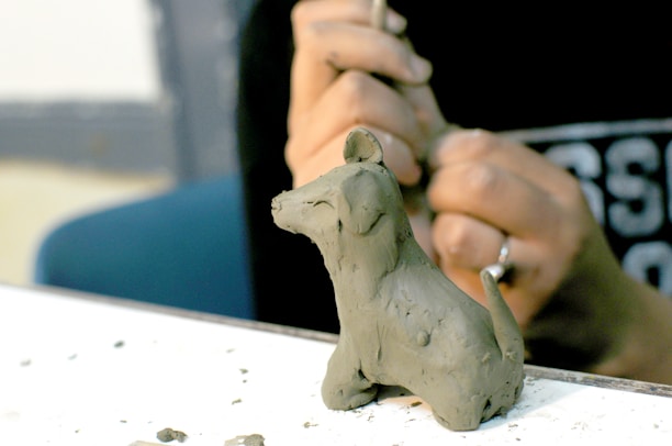 A person is sculpting a small clay figure of a dog with basic features and smooth texture. The focus is on the hands and the clay model, with tools used in sculpting visible in the background.