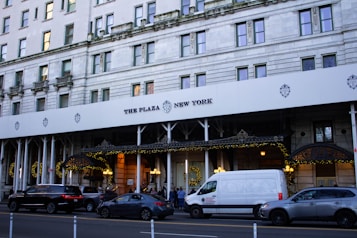 The image depicts the facade of a grand hotel with a white canopy and the words 'The Plaza New York' displayed prominently. The building features classic architectural elements with numerous windows and ornate details. Festive decorations and lights adorn the entrance area. Several vehicles, including a white van and cars, are parked along the street in front of the hotel.