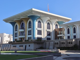 A grand architectural structure with a unique design featuring large arched windows and decorative columns. The building exhibits a blend of modern and traditional elements with geometric patterns and intricate detailing. Bright sunlight casts shadows on the fa&ccedil;ade, highlighting shades of blue, beige, and white. A well-maintained garden and courtyard with green lawns are in the foreground.