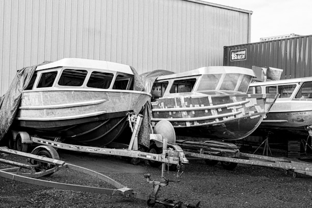 Several boats are under construction or repair. They are positioned on trailers in an industrial area, surrounded by a large warehouse and shipping containers. Protective tarps partially cover some of the boats.