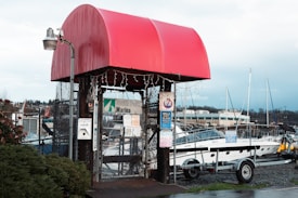 A marina entrance with a bold red awning sheltering a gated entry point. The structure is surrounded by a chain-link fence and adorned with various instructional and warning signs. There are boats visible in the background along with a few buildings and a cloudy sky.