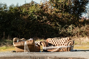 A pair of brown, tufted sofas are abandoned outdoors near a dense, green shrubbery. One sofa is fully visible, while the other is partially wrapped in plastic. The area around the sofas is covered with gravel and scattered debris, and the lighting suggests a sunny day.