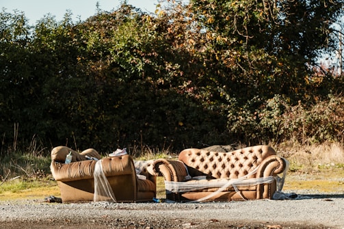 A pair of brown, tufted sofas are abandoned outdoors near a dense, green shrubbery. One sofa is fully visible, while the other is partially wrapped in plastic. The area around the sofas is covered with gravel and scattered debris, and the lighting suggests a sunny day.