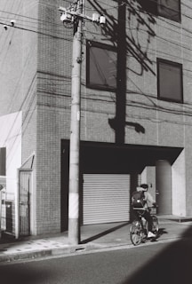 A black-and-white photograph captures a person riding a bicycle along a street. The individual is seen wearing casual clothing and carrying a backpack. The scene is set beside a large, modern building with a tile facade, featuring several windows and a large closed garage door. Shadows of utility poles and wires stretch across the wall, adding to the urban atmosphere.