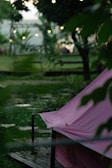 A pink tent is set up outdoors on a wooden platform surrounded by lush green foliage. Soft, ambient lighting is visible in the background, creating a serene atmosphere. The setting appears to be a garden or a park, with trees and plants adding to the natural scene.