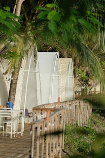 Several tent-like white structures are arranged beside a wooden boardwalk, surrounded by lush green palm trees and foliage. A small patio area with chairs and a table is visible, creating a tranquil and inviting outdoor setting.