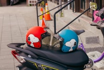 Two motorcycle helmets featuring a cartoon character design, one red and one blue, are placed on the seat of a parked scooter. In the background, there are orange traffic cones and a stairway leading to a building entrance.