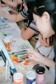 A group of people engaged in a painting activity, focusing on a table covered with various art materials. Paint tubes, brushes, and a colorful palette are prominently displayed, suggesting a creative and collaborative environment.