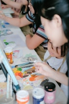 A group of people engaged in a painting activity, focusing on a table covered with various art materials. Paint tubes, brushes, and a colorful palette are prominently displayed, suggesting a creative and collaborative environment.