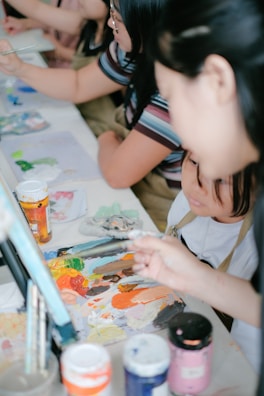 A group of people engaged in a painting activity, focusing on a table covered with various art materials. Paint tubes, brushes, and a colorful palette are prominently displayed, suggesting a creative and collaborative environment.