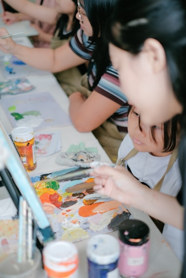 A group of people engaged in a painting activity, focusing on a table covered with various art materials. Paint tubes, brushes, and a colorful palette are prominently displayed, suggesting a creative and collaborative environment.