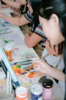 A group of people engaged in a painting activity, focusing on a table covered with various art materials. Paint tubes, brushes, and a colorful palette are prominently displayed, suggesting a creative and collaborative environment.