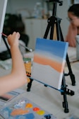 An artist is painting a canvas on an easel. The canvas depicts a gradient sky with shades of blue, orange, and white, resembling a sunset. The artist's hand is holding a brush, applying more paint. A palette with various colors is on the table, including orange, yellow, and blue. In the background, a person appears to be looking at the artwork.