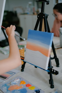 An artist is painting a canvas on an easel. The canvas depicts a gradient sky with shades of blue, orange, and white, resembling a sunset. The artist's hand is holding a brush, applying more paint. A palette with various colors is on the table, including orange, yellow, and blue. In the background, a person appears to be looking at the artwork.