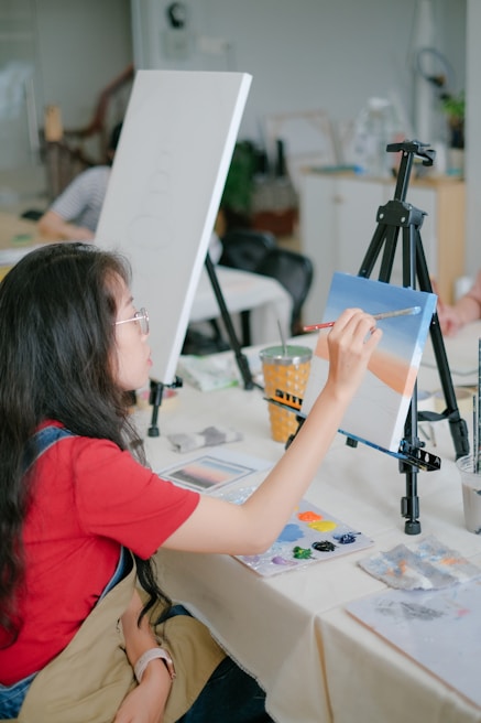 A person with long hair, wearing a red shirt and apron, is painting on a canvas set on an easel. The scene is indoors, likely a classroom or studio setting. The person has a brush in hand, applying light colors to the canvas. Art supplies and paint palettes are visible on the table.