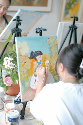 A young woman is painting on a canvas set on an easel. The artwork features a girl in a white dress with colorful designs and a background of yellow and blue hues. Nearby, art supplies like brushes and paint jars are visible on the table, and a potted plant with flowers adds a touch of nature.