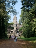A large stone temple structure with intricate carvings and a tall central spire is surrounded by lush greenery and trees. A crowd of people is walking along a leaf-strewn path towards the entrance, suggesting a tourist destination.