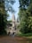 A large stone temple structure with intricate carvings and a tall central spire is surrounded by lush greenery and trees. A crowd of people is walking along a leaf-strewn path towards the entrance, suggesting a tourist destination.