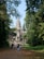 A large stone temple structure with intricate carvings and a tall central spire is surrounded by lush greenery and trees. A crowd of people is walking along a leaf-strewn path towards the entrance, suggesting a tourist destination.