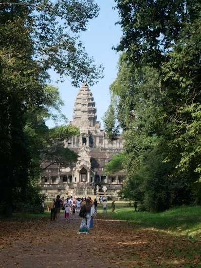 A large stone temple structure with intricate carvings and a tall central spire is surrounded by lush greenery and trees. A crowd of people is walking along a leaf-strewn path towards the entrance, suggesting a tourist destination.