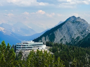 A modern building with a large deck is nestled among green pine trees in a mountainous area. The structure is surrounded by a vast expanse of mountains under a partly cloudy sky, creating a picturesque and serene atmosphere.