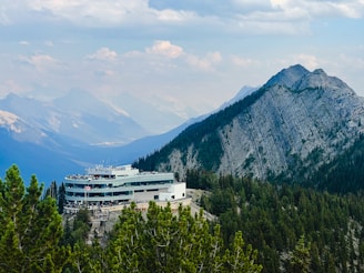 A modern building with a large deck is nestled among green pine trees in a mountainous area. The structure is surrounded by a vast expanse of mountains under a partly cloudy sky, creating a picturesque and serene atmosphere.