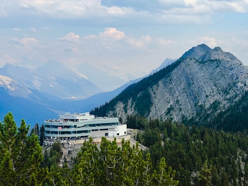 A modern building with a large deck is nestled among green pine trees in a mountainous area. The structure is surrounded by a vast expanse of mountains under a partly cloudy sky, creating a picturesque and serene atmosphere.