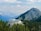 A modern building with a large deck is nestled among green pine trees in a mountainous area. The structure is surrounded by a vast expanse of mountains under a partly cloudy sky, creating a picturesque and serene atmosphere.