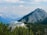 A modern building with a large deck is nestled among green pine trees in a mountainous area. The structure is surrounded by a vast expanse of mountains under a partly cloudy sky, creating a picturesque and serene atmosphere.