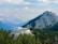 A modern building with a large deck is nestled among green pine trees in a mountainous area. The structure is surrounded by a vast expanse of mountains under a partly cloudy sky, creating a picturesque and serene atmosphere.