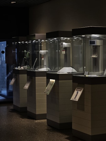 Several illuminated display cases stand in a dimly lit environment. Each case contains scientific or technological instruments, possibly museum exhibits. Informational plaques are attached to the front of each pedestal.