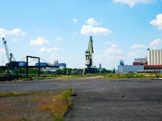 An industrial landscape featuring large cranes and warehouses against a backdrop of a bright blue sky with scattered clouds. The foreground displays an open, somewhat barren space with some vegetation lining the edges. There are buildings and structures visible at a distance, along with some trees.