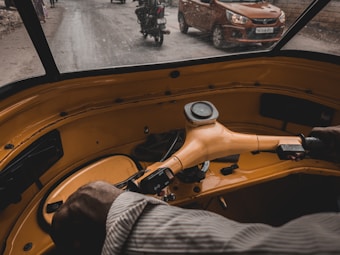 The interior view of an auto rickshaw with a person's hands on the steering bar. Visible through the windshield, there is a car and a scooter on the road, with buildings lining the street.
