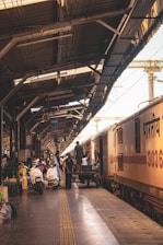 A busy train station platform with people engaged in various activities. A train is stationed on the right side, with individuals loading and unloading luggage. The platform extends into the distance, covered by a metal roof with beams and cables. Various luggage items, bicycles, and boxes are strewn around the platform, indicating an active scene.
