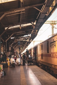 A busy train station platform with people engaged in various activities. A train is stationed on the right side, with individuals loading and unloading luggage. The platform extends into the distance, covered by a metal roof with beams and cables. Various luggage items, bicycles, and boxes are strewn around the platform, indicating an active scene.