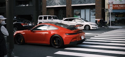 A red sports car is stopped at a zebra crossing in an urban street, surrounded by a white SUV and a white sedan. A person wearing a mask stands on one side of the crossing, while another person on a bicycle is visible near a building with signage in Japanese. The background features buildings with brick and glass exteriors.