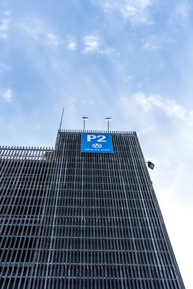 A multi-story parking structure with metal grid facade rising towards a bright sky. A blue sign labeled 'P2' is prominently displayed on the building, indicating a parking level.