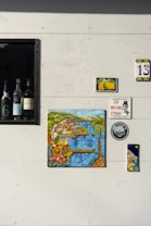 A white wall adorned with various decorative items including ceramic tiles with colorful illustrations. A framed section contains several wine bottles. The tiles feature vibrant images of Italian coastal scenery and lemons, with one labeled with the number 13. A small plaque displays the phrase 'CCA NISCIUNO E' FESSO' next to a circular logo.