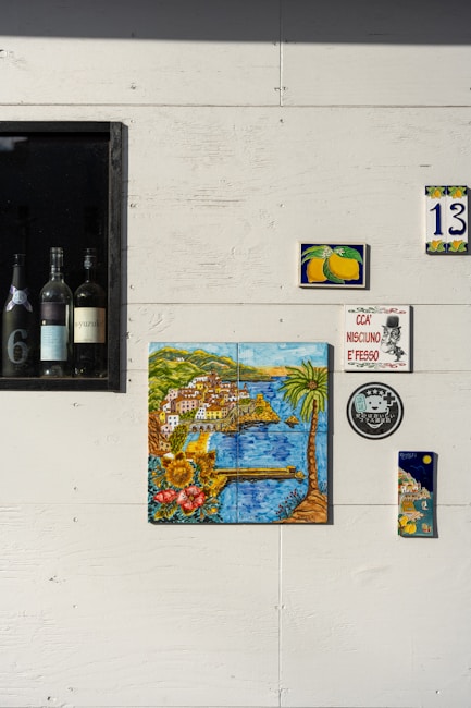 A white wall adorned with various decorative items including ceramic tiles with colorful illustrations. A framed section contains several wine bottles. The tiles feature vibrant images of Italian coastal scenery and lemons, with one labeled with the number 13. A small plaque displays the phrase 'CCA NISCIUNO E' FESSO' next to a circular logo.