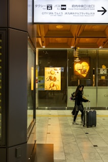 A person walks through a modern indoor space, pulling a suitcase. The surroundings feature commercial signage, including a Godiva chocolate display, and overhead directional signs in both Japanese and English for facilities such as a taxi or bus station and a spa.