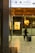 A person walks through a modern indoor space, pulling a suitcase. The surroundings feature commercial signage, including a Godiva chocolate display, and overhead directional signs in both Japanese and English for facilities such as a taxi or bus station and a spa.