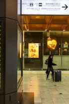 A person walks through a modern indoor space, pulling a suitcase. The surroundings feature commercial signage, including a Godiva chocolate display, and overhead directional signs in both Japanese and English for facilities such as a taxi or bus station and a spa.