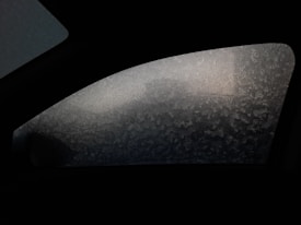 A car window covered in frost patterns, partially obscuring the view of the outside, with variations of light penetrating through the ice crystals.